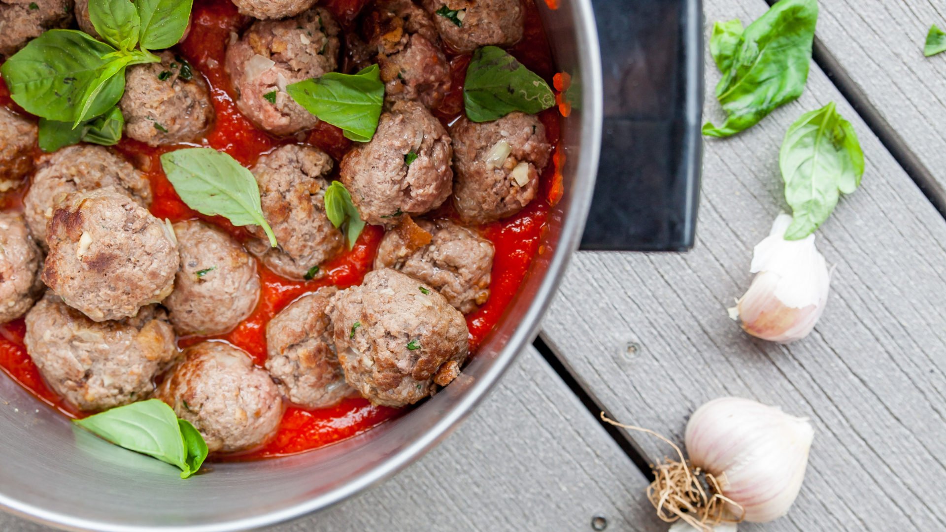 meatballs in a pan, basil leaves and garlic heads beside