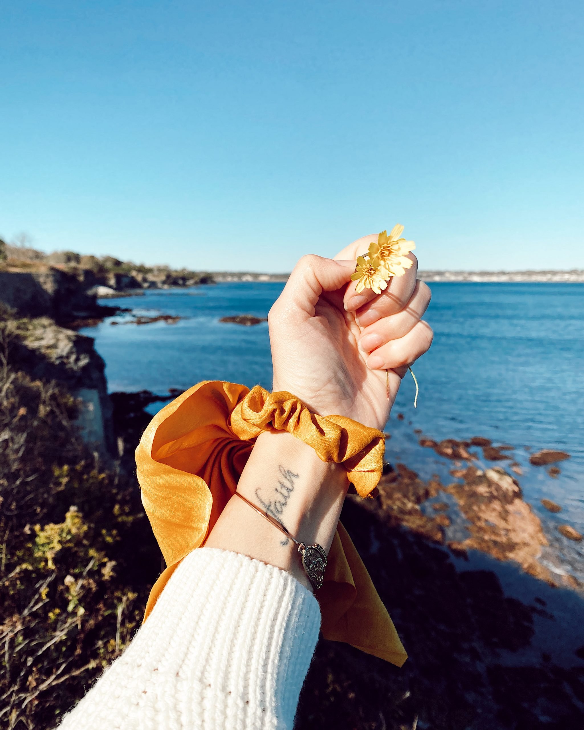 a women's hand holding a daisy wearing a yellow scrunchie with the ocean in the background.