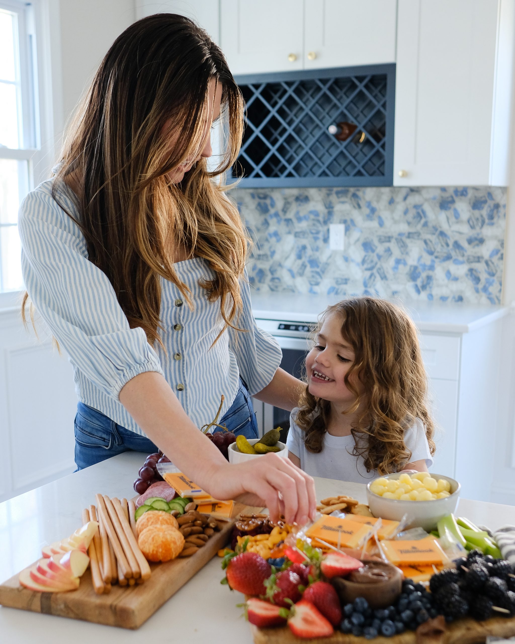 a woman and her daughter both with long brown hair eating from a snack board