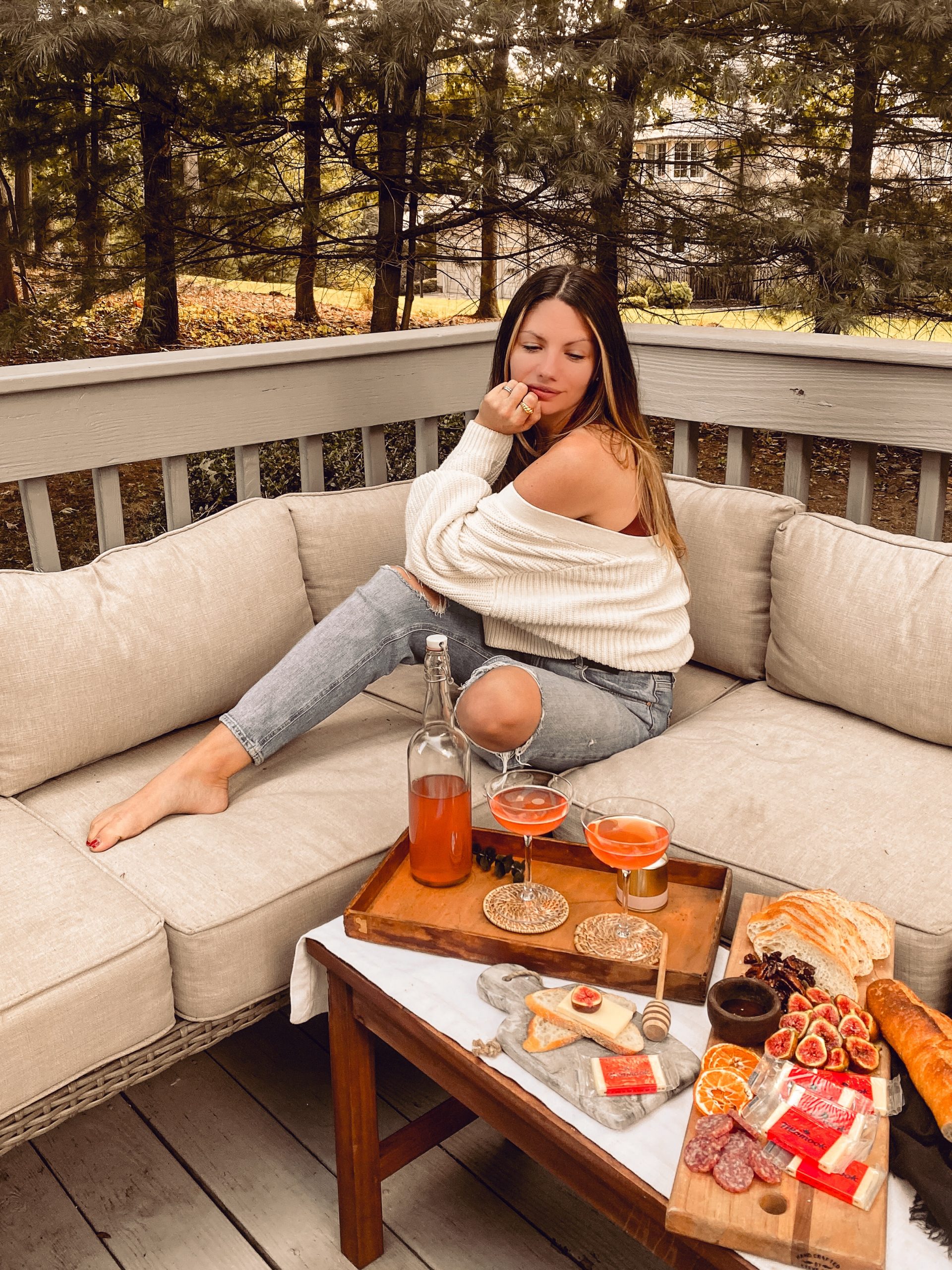 a woman sits on patio furniture with long brown hair and a white sweater in front of a large cheese plate. 